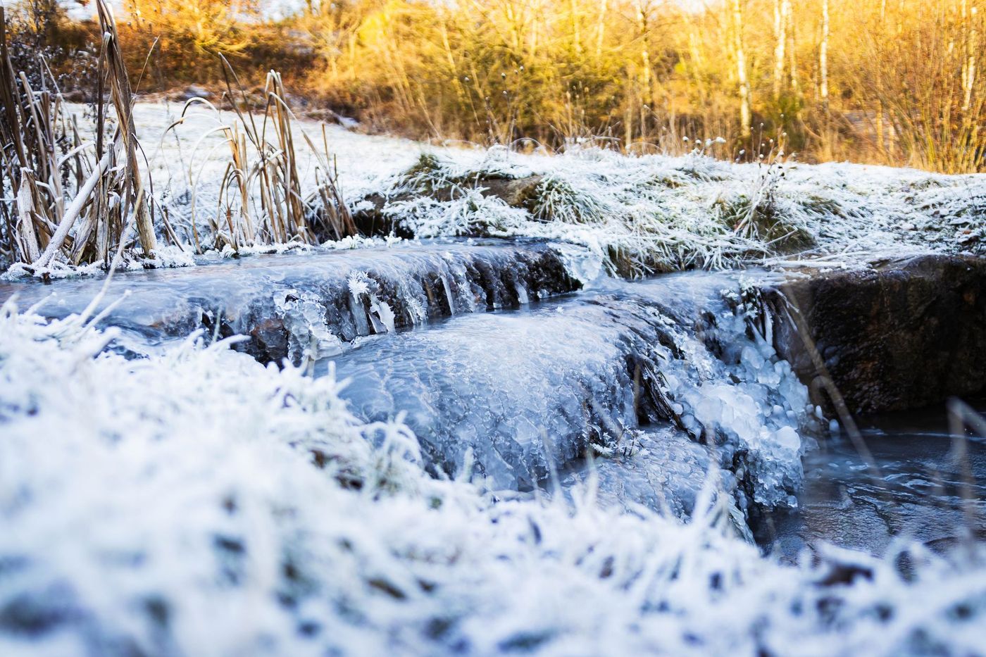 A close-up of a small, partially frozen stream or waterfall in a snowy, natural outdoor setting.