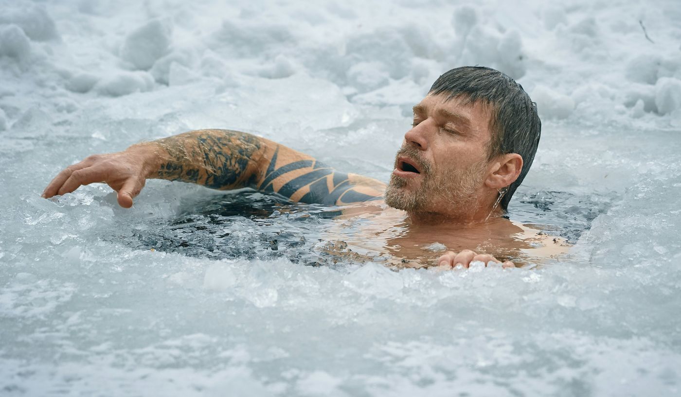 A man with a tattooed arm takes a cold plunge in a hole cut into a frozen, snow-covered body of water outdoors.