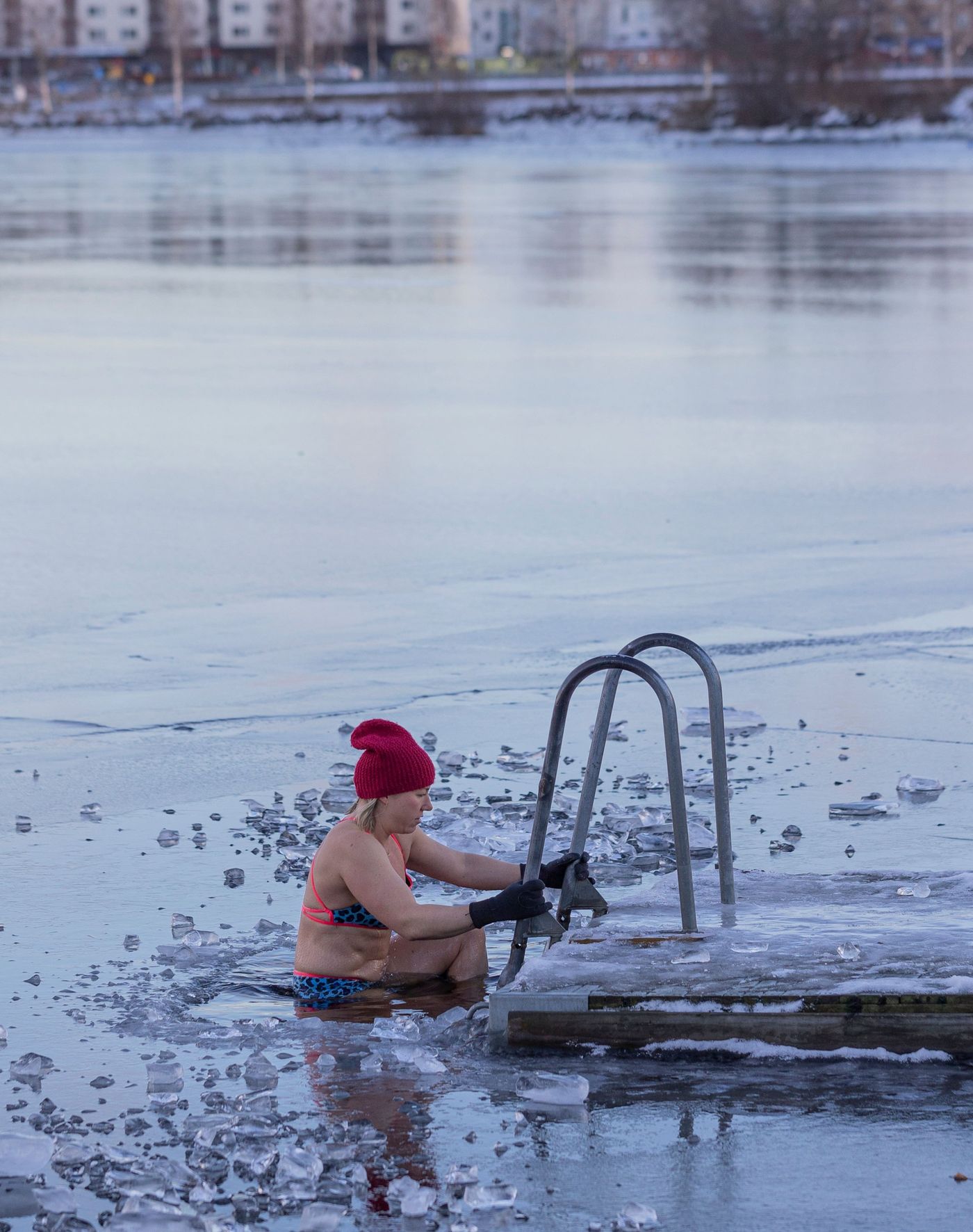 A woman in a bikini and beanie is in an icy lake or sea, holding onto a ladder attached to a dock.