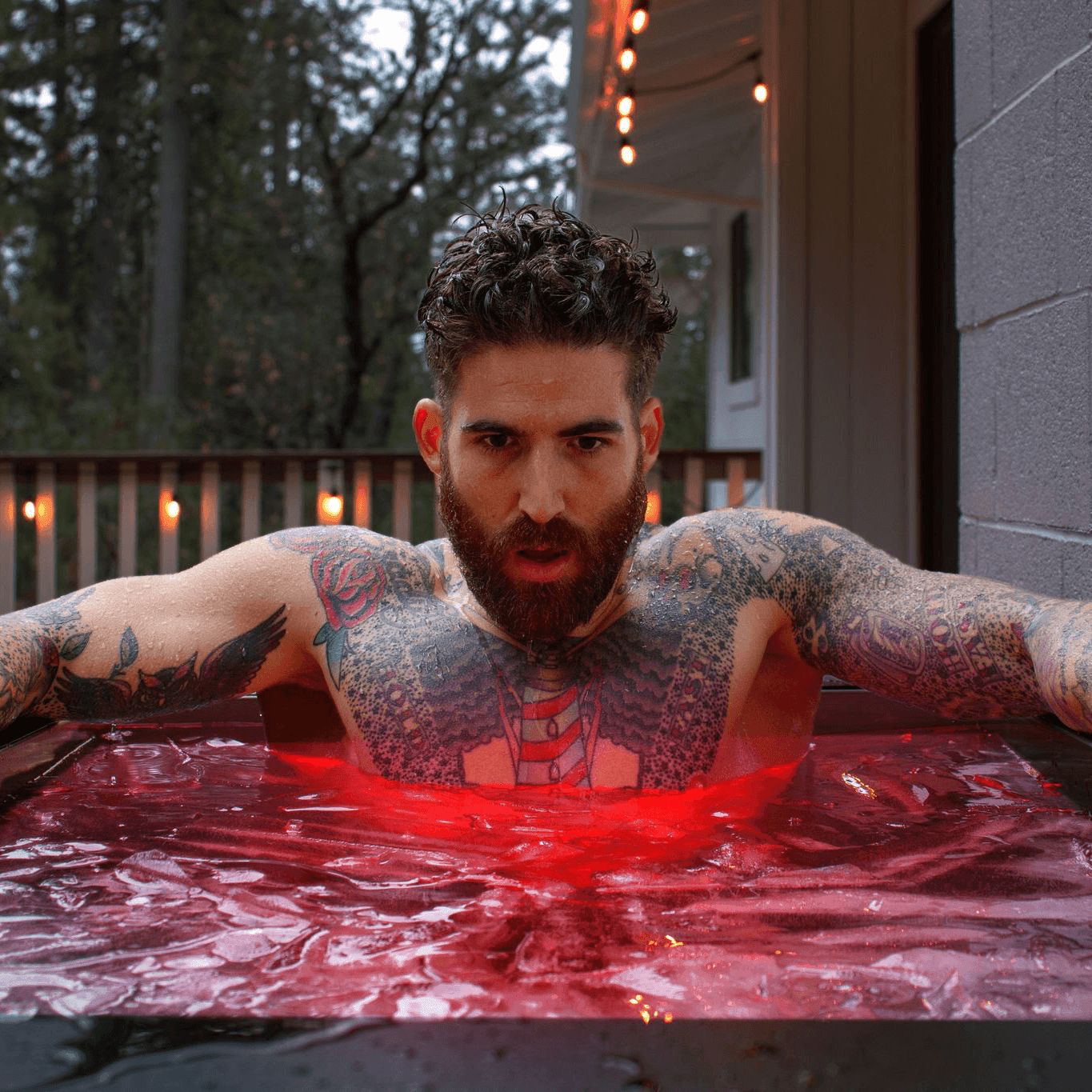 A shirtless, tattooed man with a beard submerged up to his chest in an outdoor cold plunge tub with red lighting.