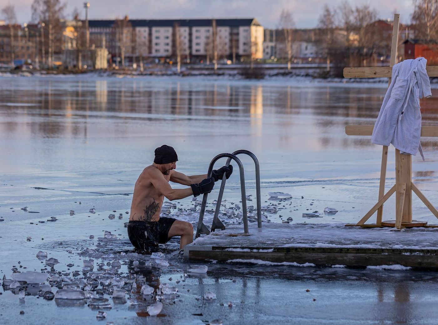 A man in shorts, a beanie, and gloves lowers himself into a hole cut in a frozen lake, holding onto a metal ladder.