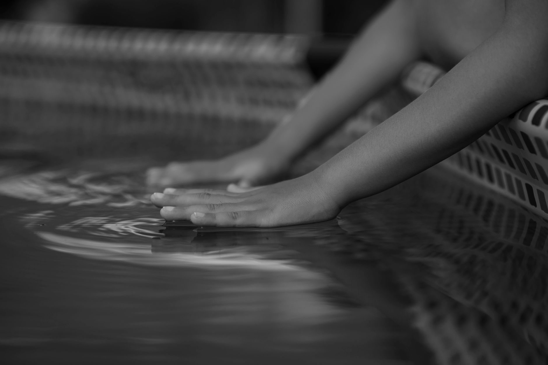 A black and white close-up of a person's hands and forearms resting on the surface of the water at the edge of a pool or tub.