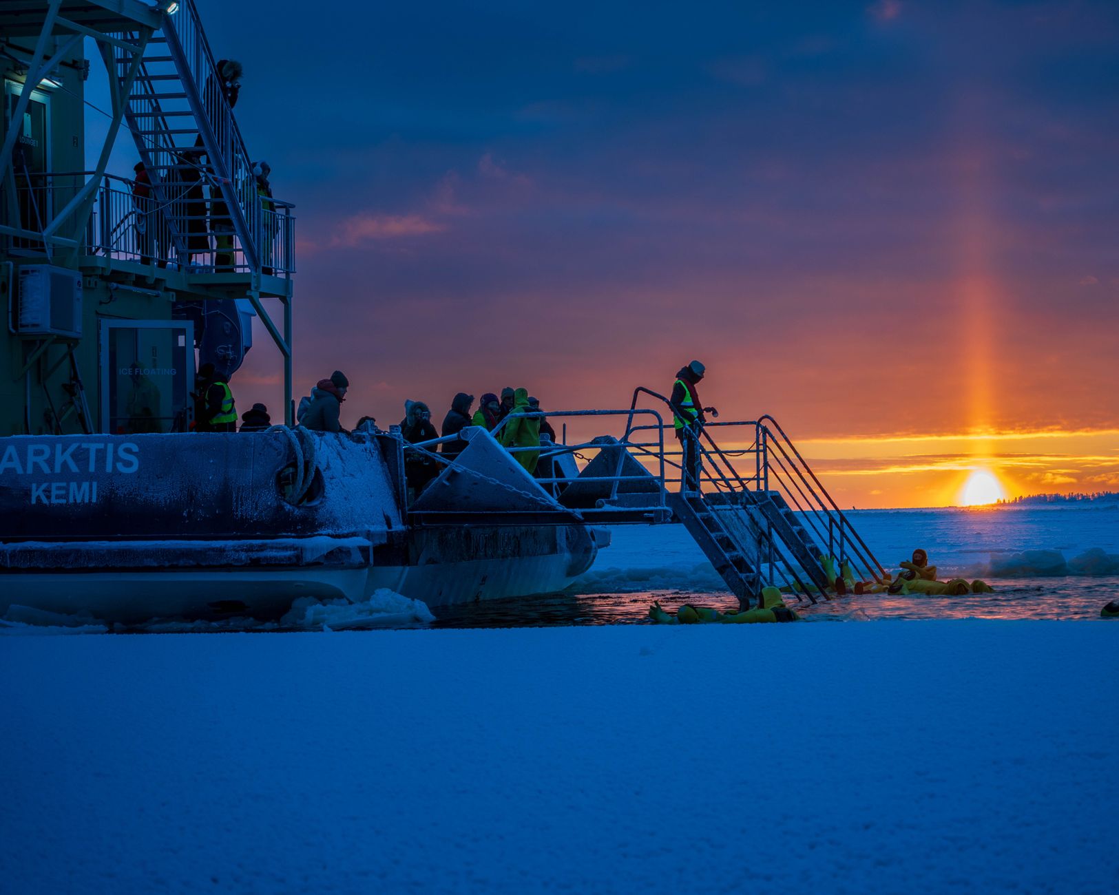People in yellow survival suits float in icy water next to a large boat at sunset in a snowy landscape.