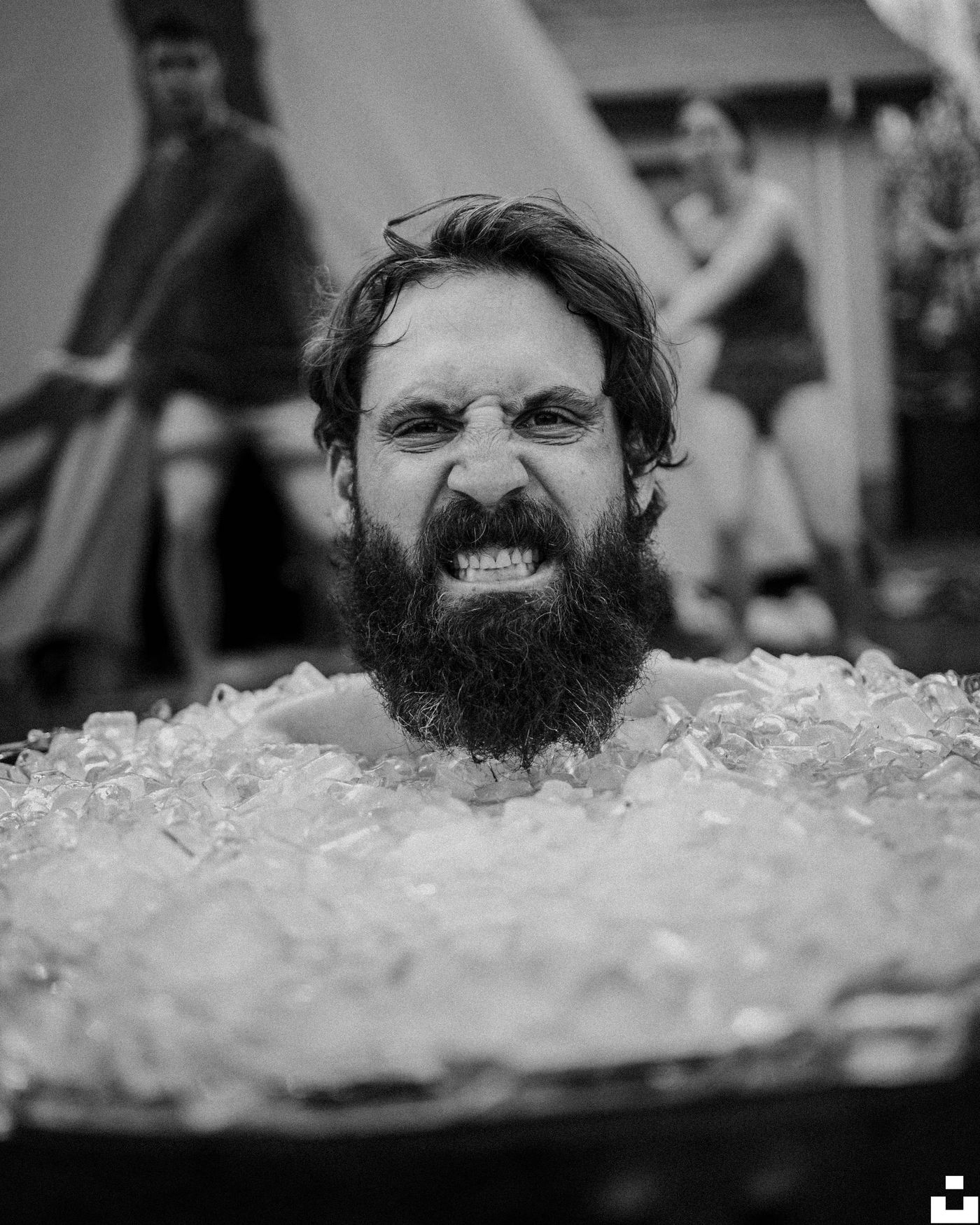 A black and white photo of a bearded man grimacing while submerged up to his neck in a tub full of ice cubes outdoors.