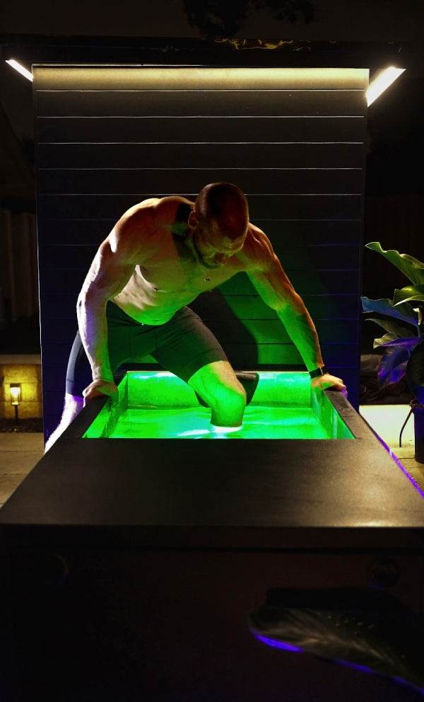 A muscular man is getting into a cold plunge tub filled with glowing green water at night.