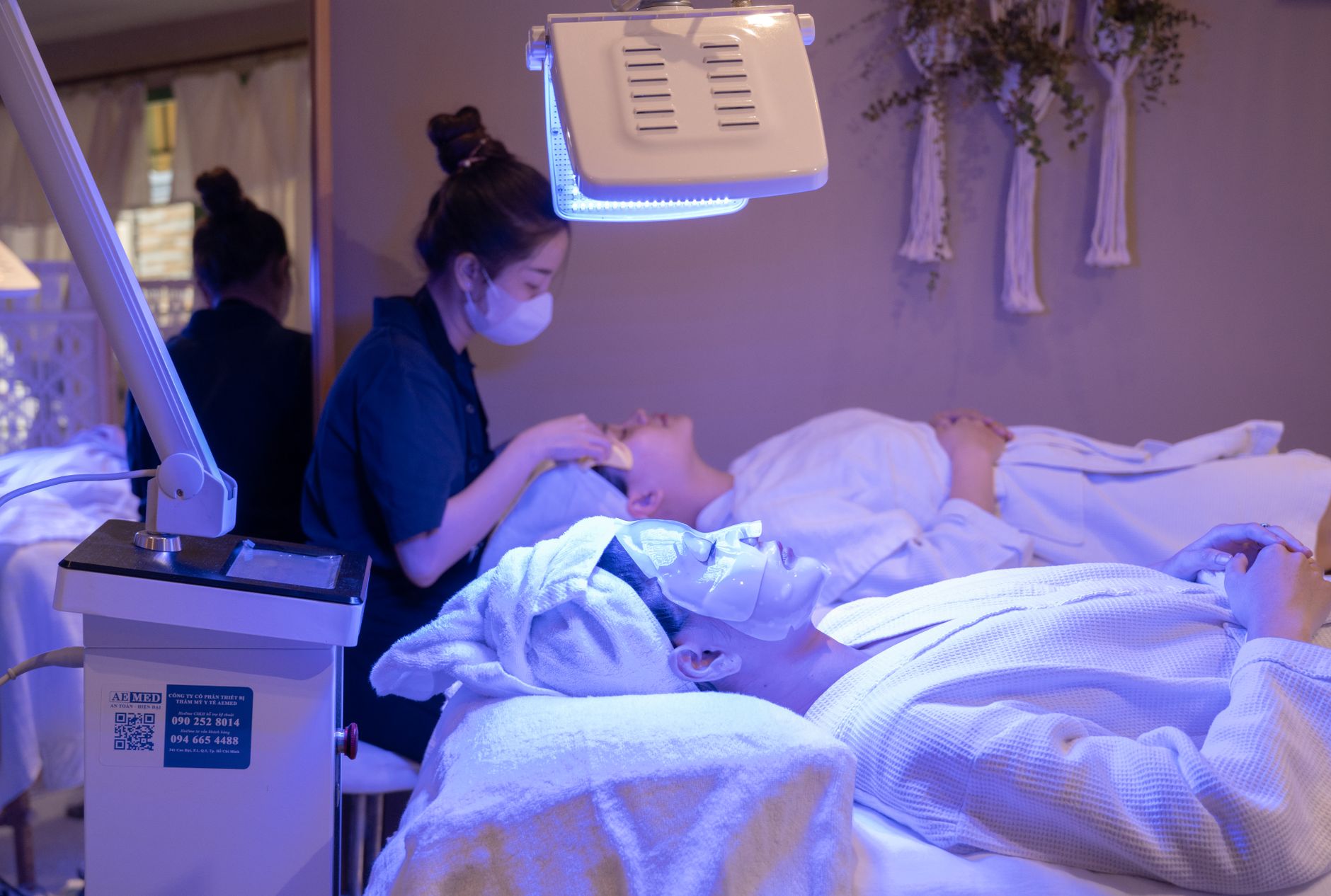 A client lies on a spa treatment bed under an overhead LED light-therapy panel while an esthetician works nearby.