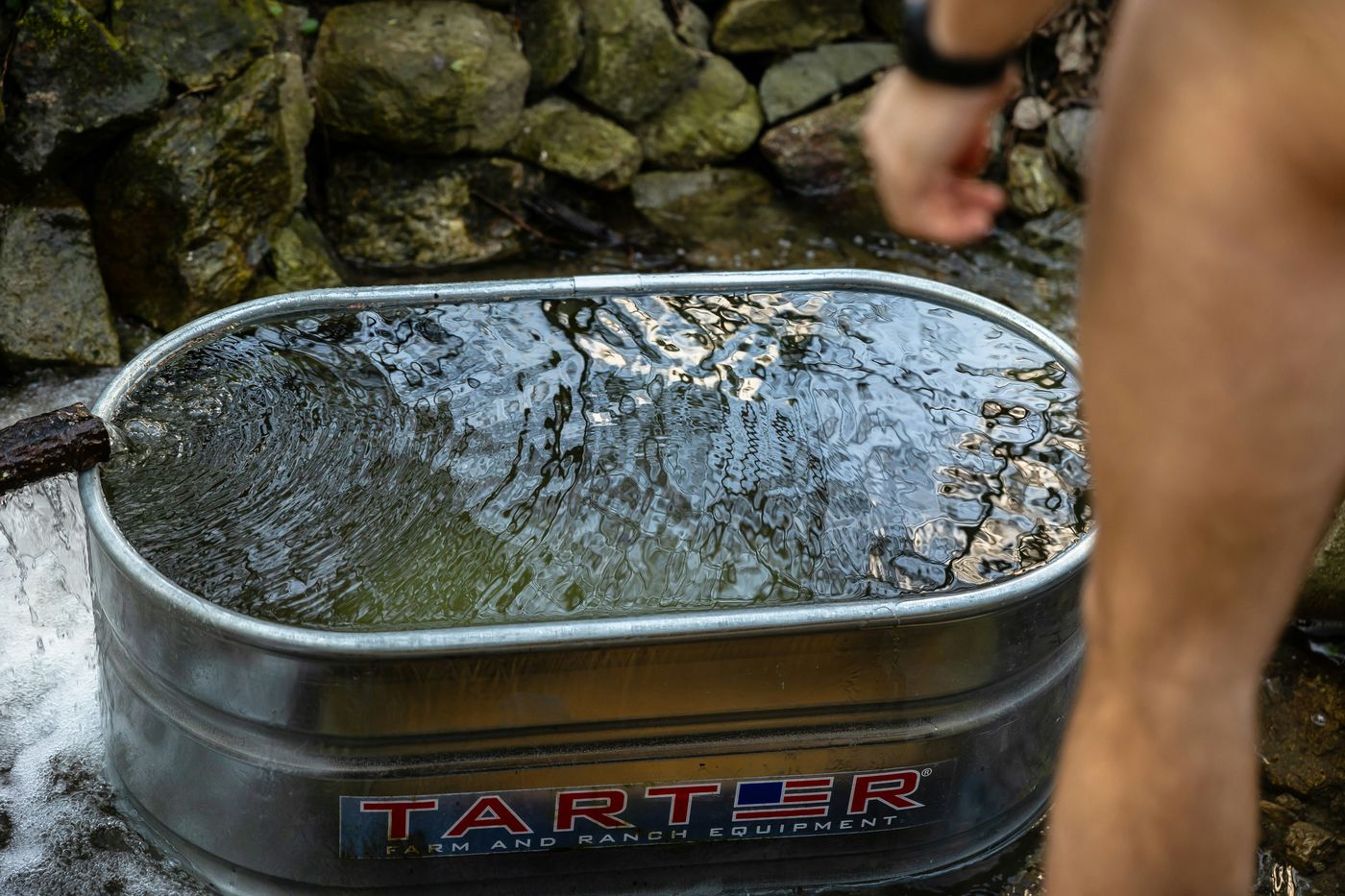 A metal farm trough is being filled with water from a spout in a rocky stream, with a person's arm and leg visible.