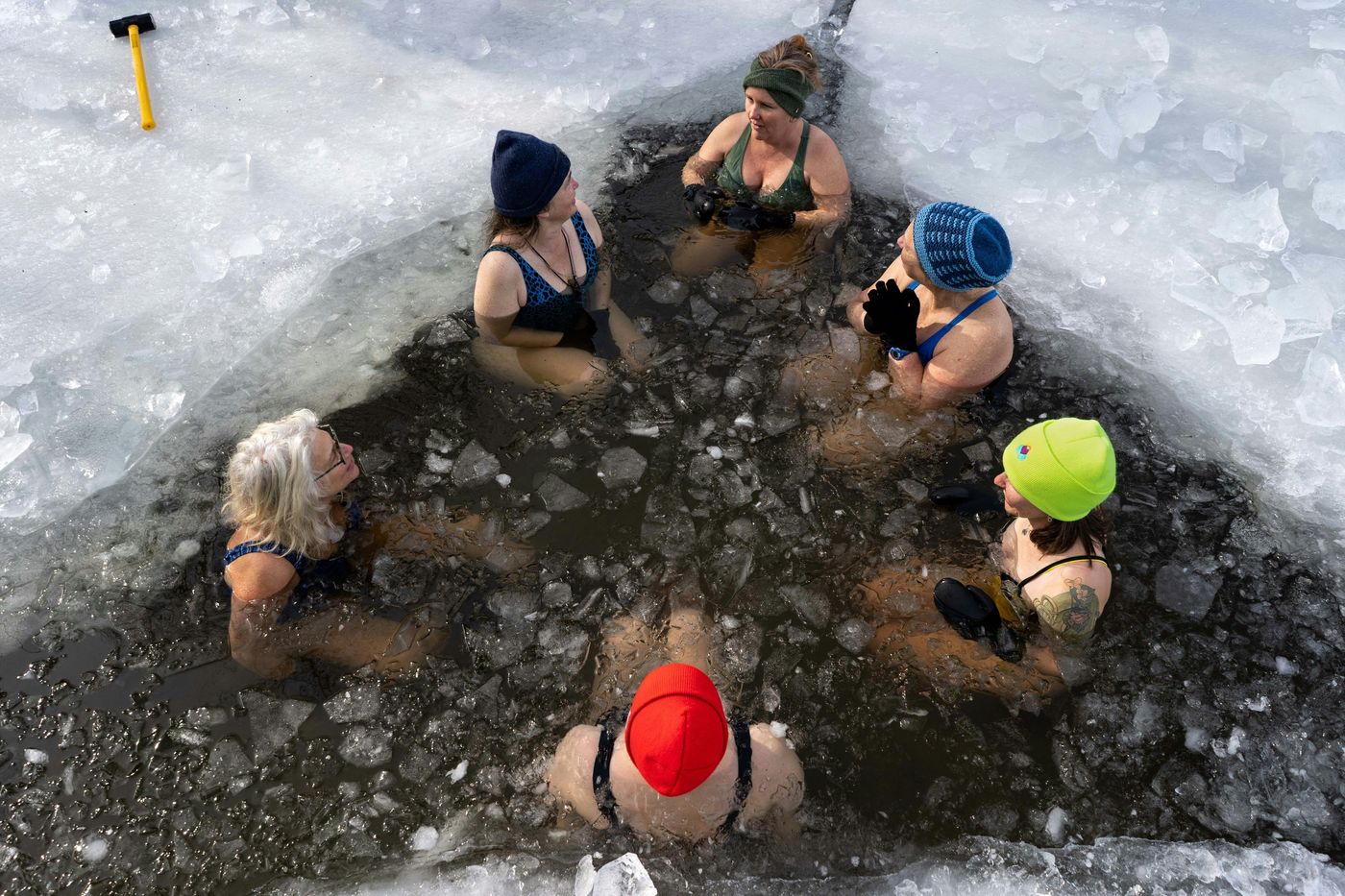 A group of women in swimsuits and hats sit in a hole cut into a frozen lake or river, taking an ice bath.